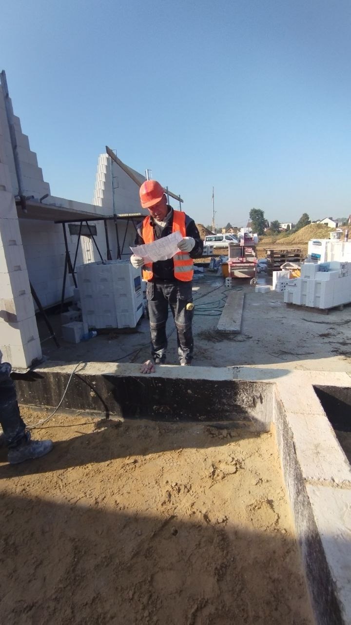 Pracownik w kasku i kamizelce odblaskowej analizuje plan na placu budowy domu z widocznymi elementami konstrukcyjnymi i zapasem bloczków.