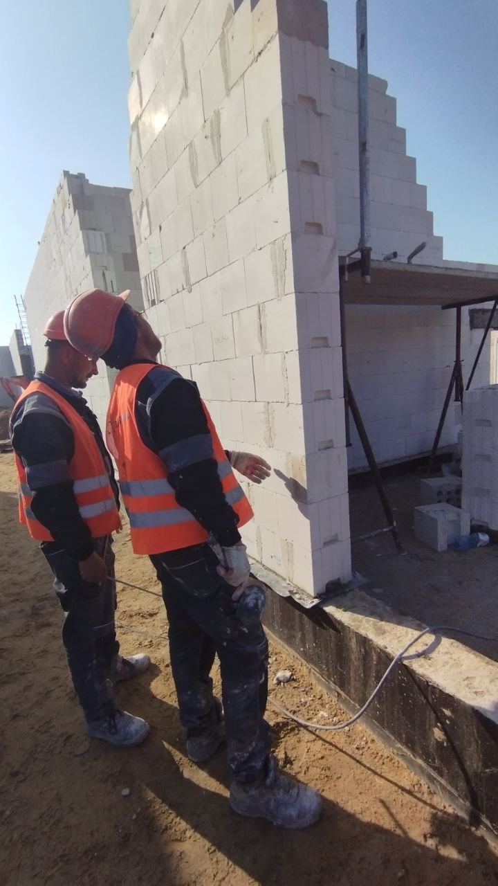 Dwóch pracowników w pomarańczowych kamizelkach i kaskach sprawdza pionowość ściany z białych bloczków, widoczne rusztowanie i kolejne ściany w tle.