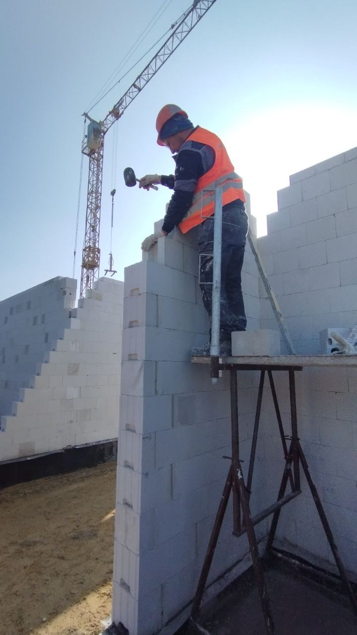 Pracownik w pomarańczowej kamizelce i kasku wyrównuje bloczki z betonu komórkowego przy użyciu gumowego młotka, widoczny dźwig budowlany w tle.