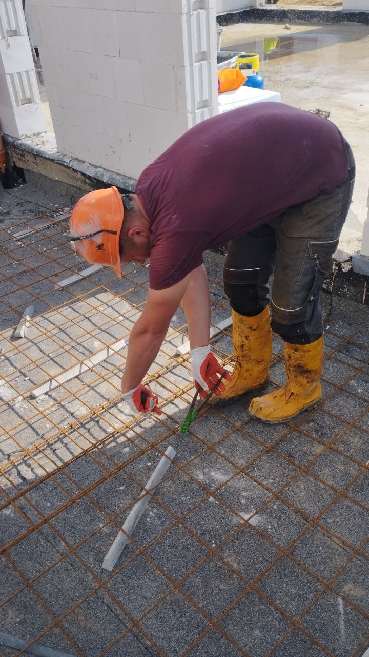 Pracownik w pomarańczowym kasku i żółtych gumowcach, zginający drut zbrojeniowy na przygotowanym fundamencie pod budowę, widoczne białe bloczki.