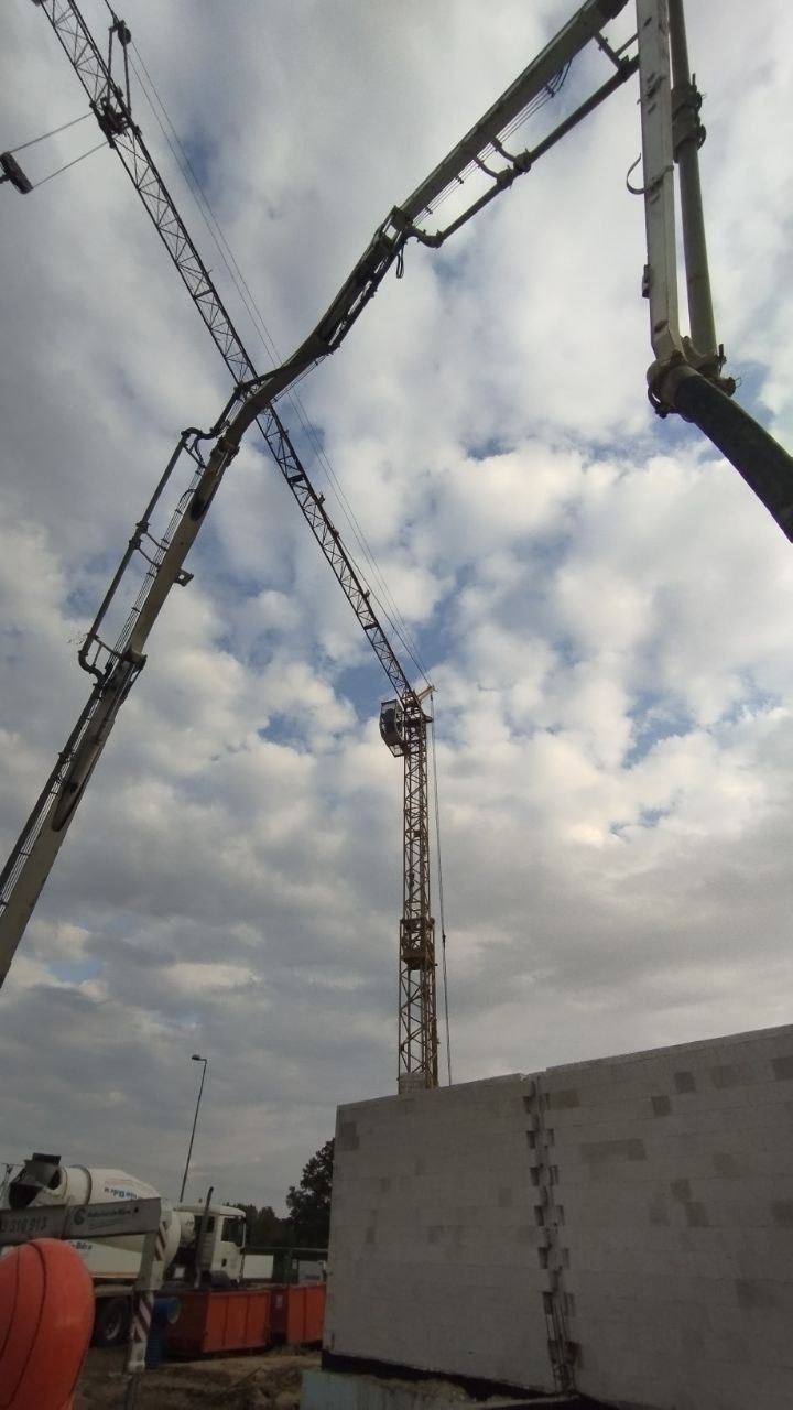 Wznoszenie ścian z bloczków na placu budowy, widoczny żuraw budowlany i pompa do betonu na tle pochmurnego nieba.