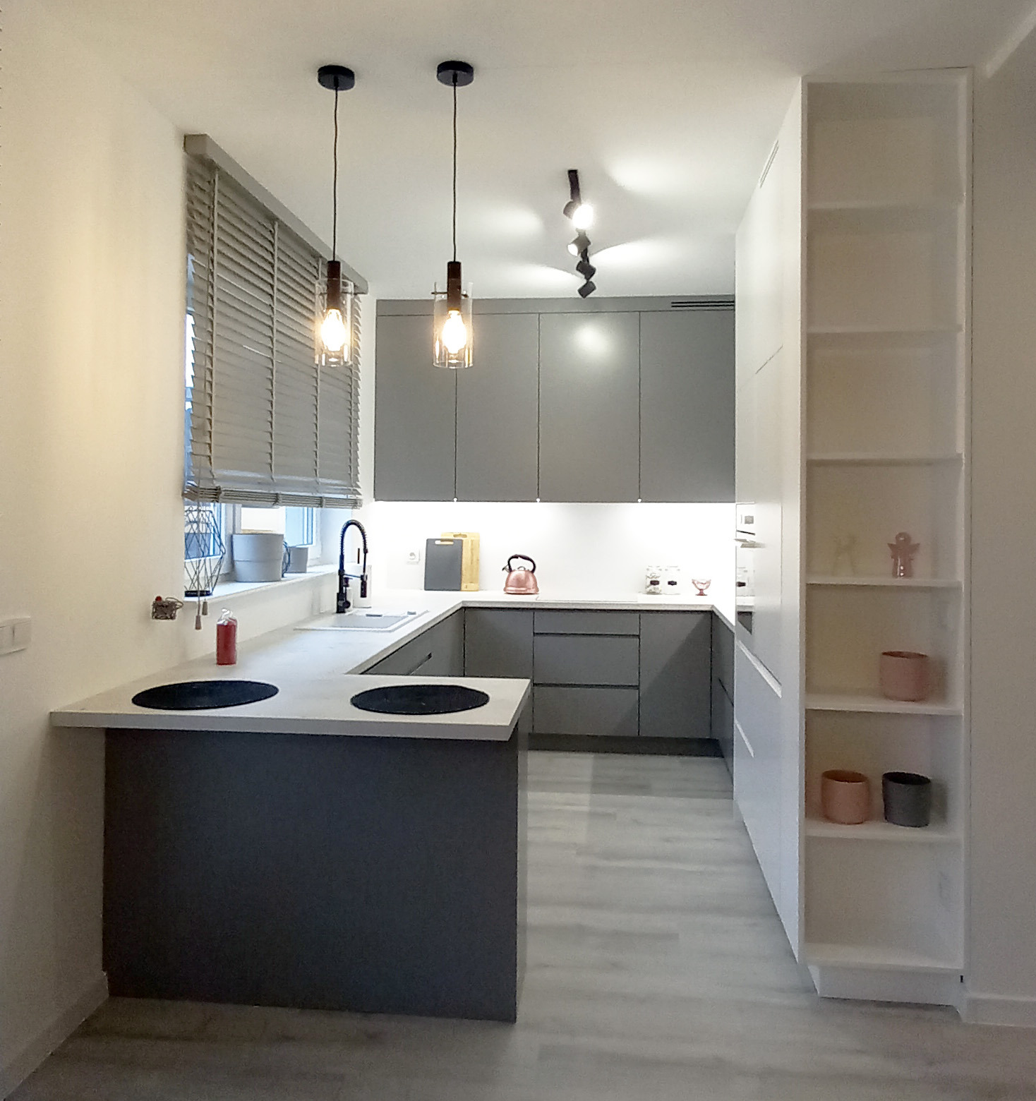 Nowoczesna kuchnia w odcieniach szarości z drewnianymi żaluzjami i minimalistycznym oświetleniem punktowym i wiszącym. Widoczna zabudowa kuchenna z frontami bez uchwytów, biały blat i wyspa z płytą...