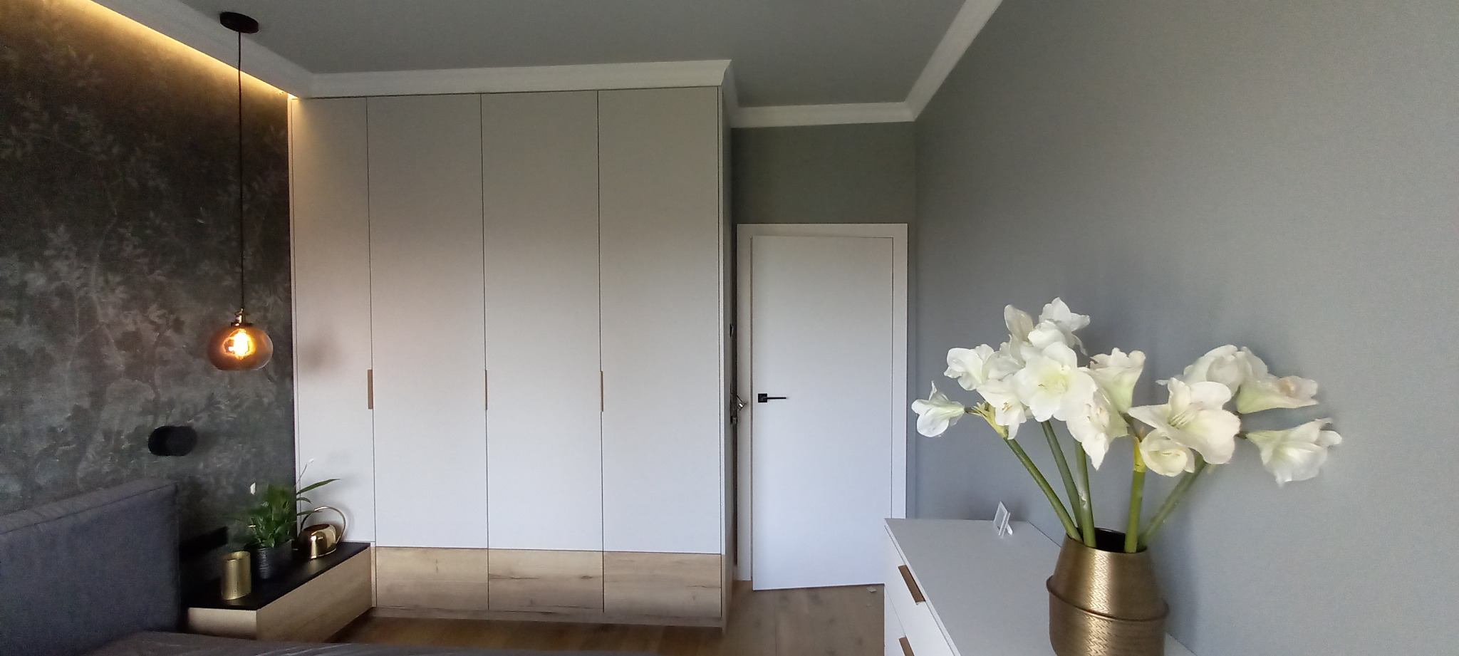 Jasna szafa wnękowa z drewnianą podstawą w sypialni z szarą ścianą i dekoracyjną lampą wiszącą.