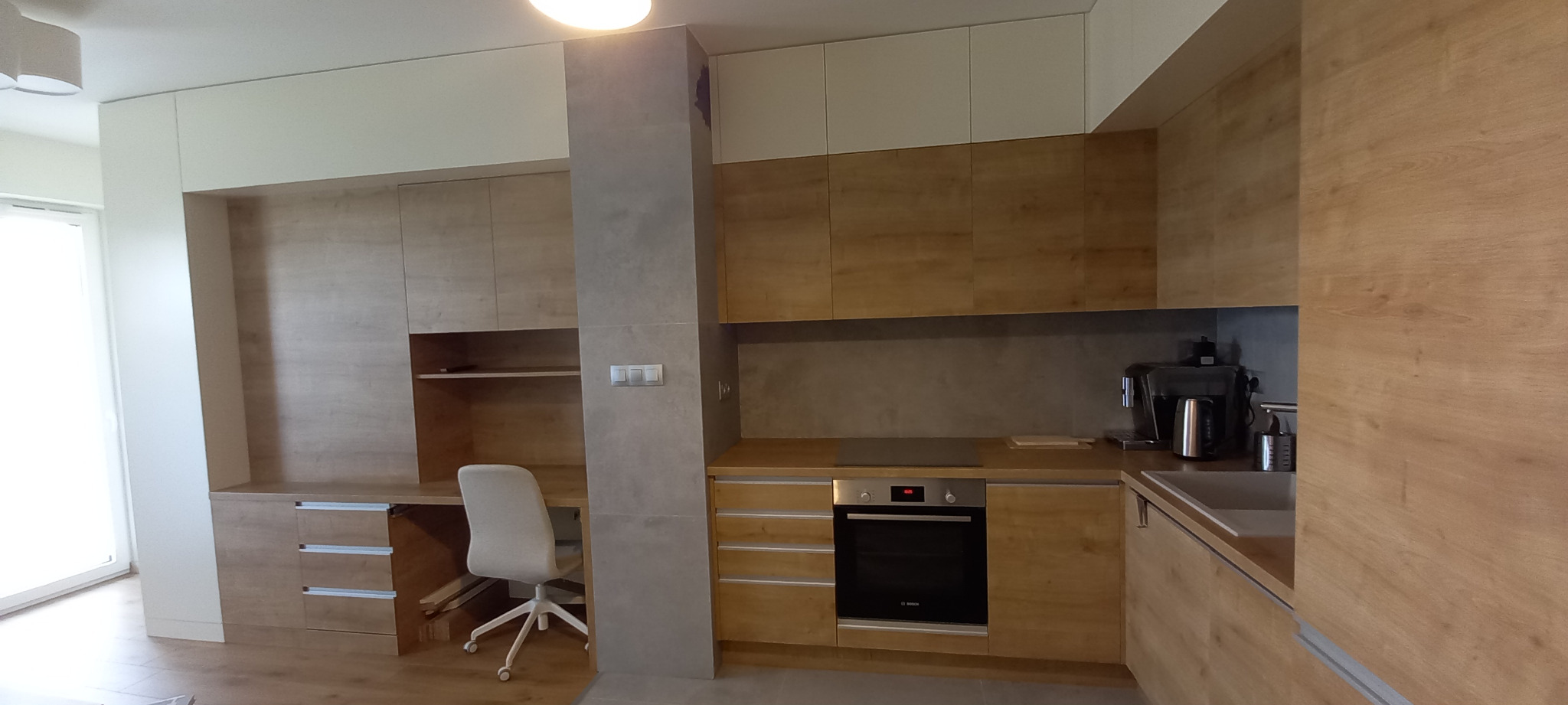 Nowoczesna kuchnia z drewnianymi frontami szafek, blatem i zabudowanym biurkiem z krzesłem na kółkach, oświetlenie sufitowe.