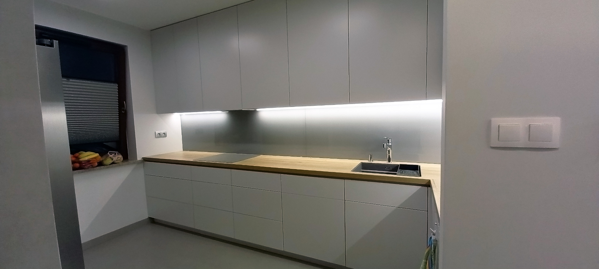 Jasna kuchnia z drewnianym blatem, oświetleniem podszafkowym LED, zlewem i płytą grzewczą, widok na okno z owocami.