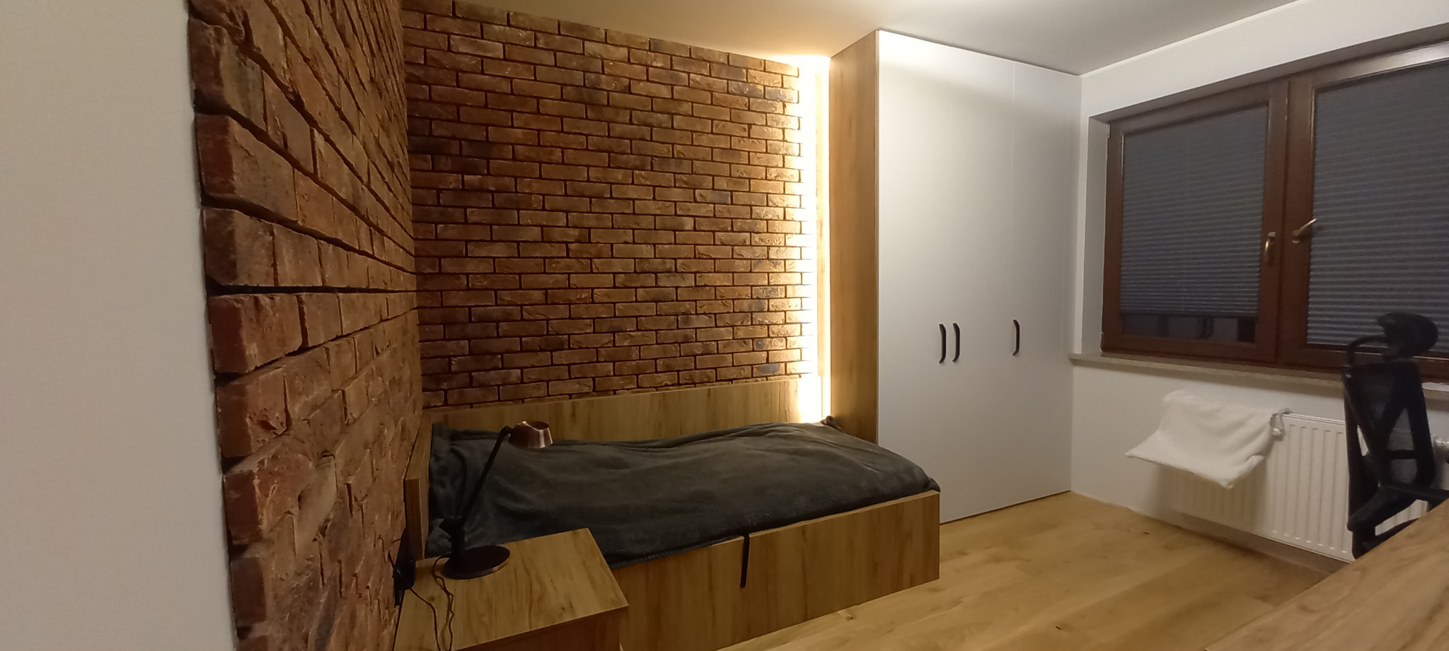 Sypialnia z łóżkiem w zabudowie z drewnianą ramą i ceglaną ścianą, oświetlona listwą LED, obok biała szafa wnękowa.