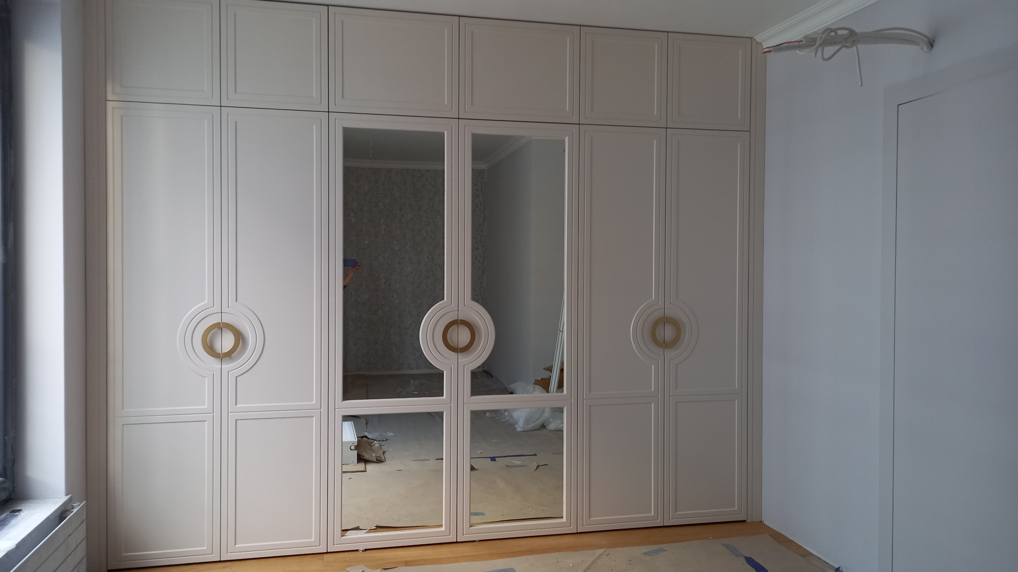 Duża, biała szafa z lustrzanymi drzwiami i okrągłymi, złotymi uchwytami, zabudowana od podłogi do sufitu, widoczna w trakcie montażu, z niezakrytym okablowaniem na suficie.