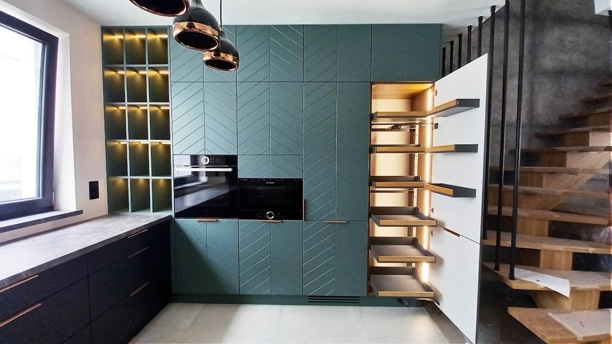 Nowoczesna kuchnia z zielonymi szafkami z wzorem jodełki, czarnymi urządzeniami AGD i wysuwaną spiżarnią z oświetleniem LED, widoczne schody w tle.