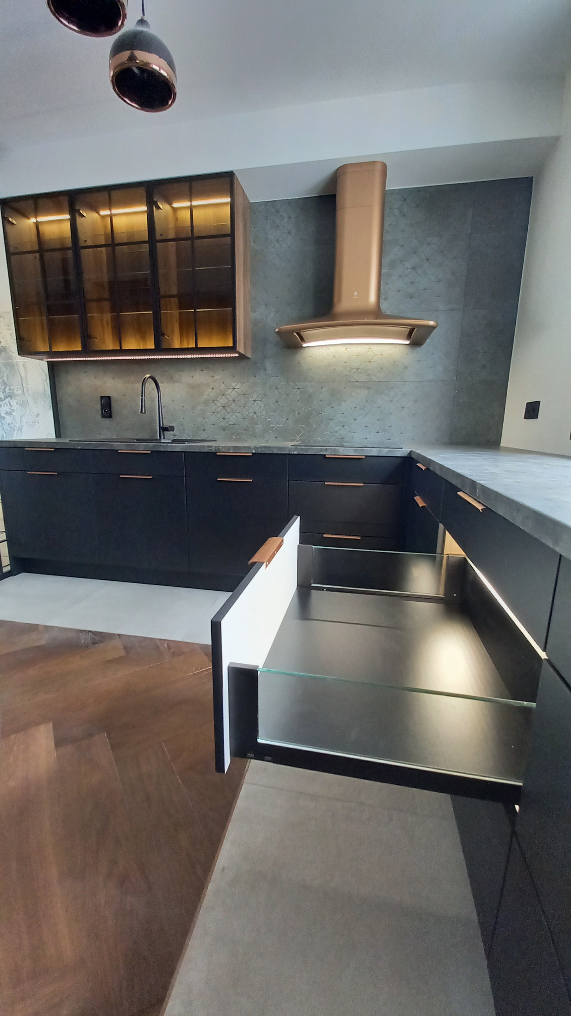 Nowoczesna kuchnia z czarnymi frontami i miedzianymi akcentami, szklana półka w otwartej szufladzie z podświetleniem LED, widoczny fragment drewnianej podłogi.