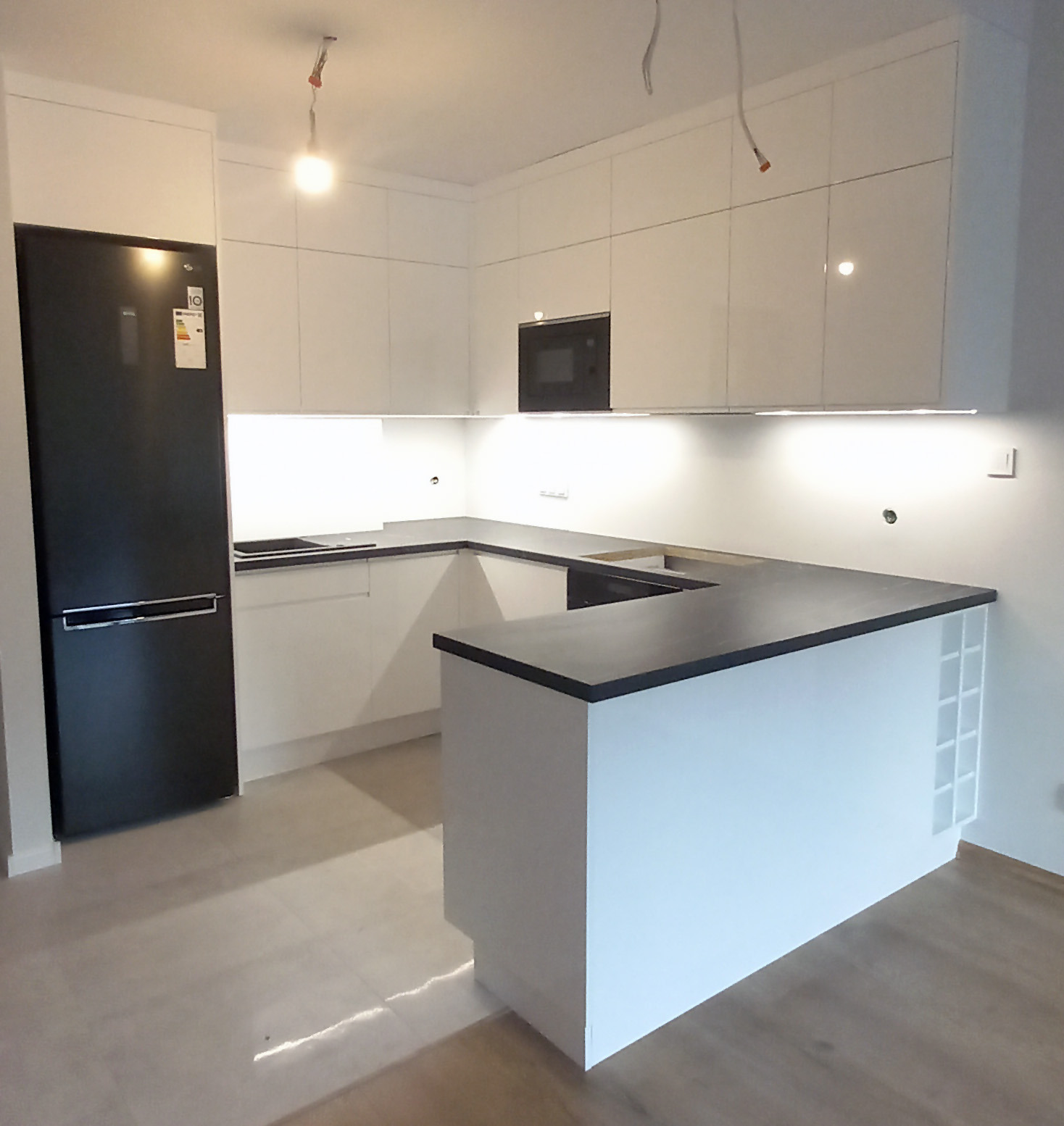 Nowoczesna, minimalistyczna kuchnia z białymi frontami szafek i czarnym blatem, oświetleniem LED pod szafkami wiszącymi i czarną lodówką po lewej stronie