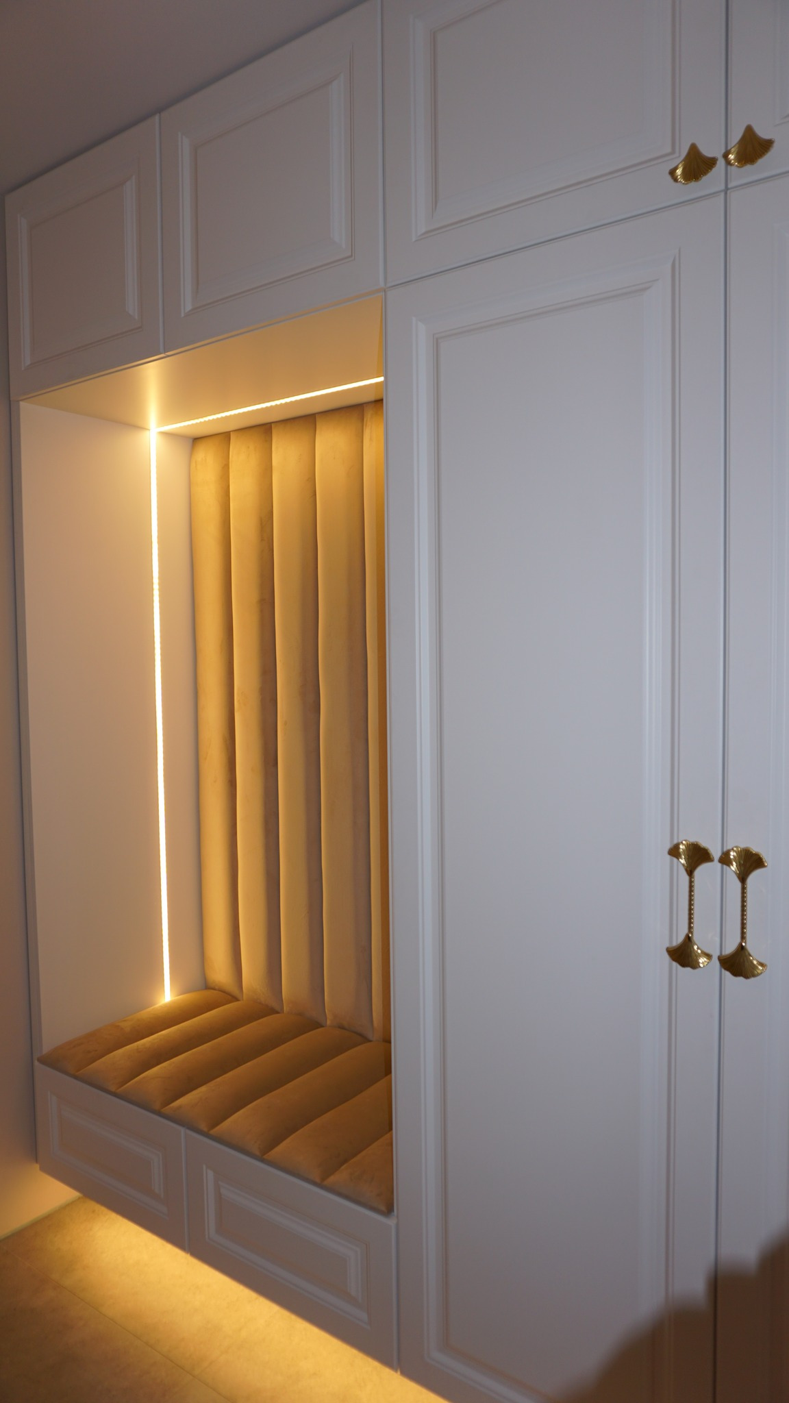 Biała zabudowa meblowa z wnęką wyłożoną tapicerowanym siedziskiem i oparciem w odcieniach beżu, oświetloną listwą LED, widoczne złote uchwyty szafek.