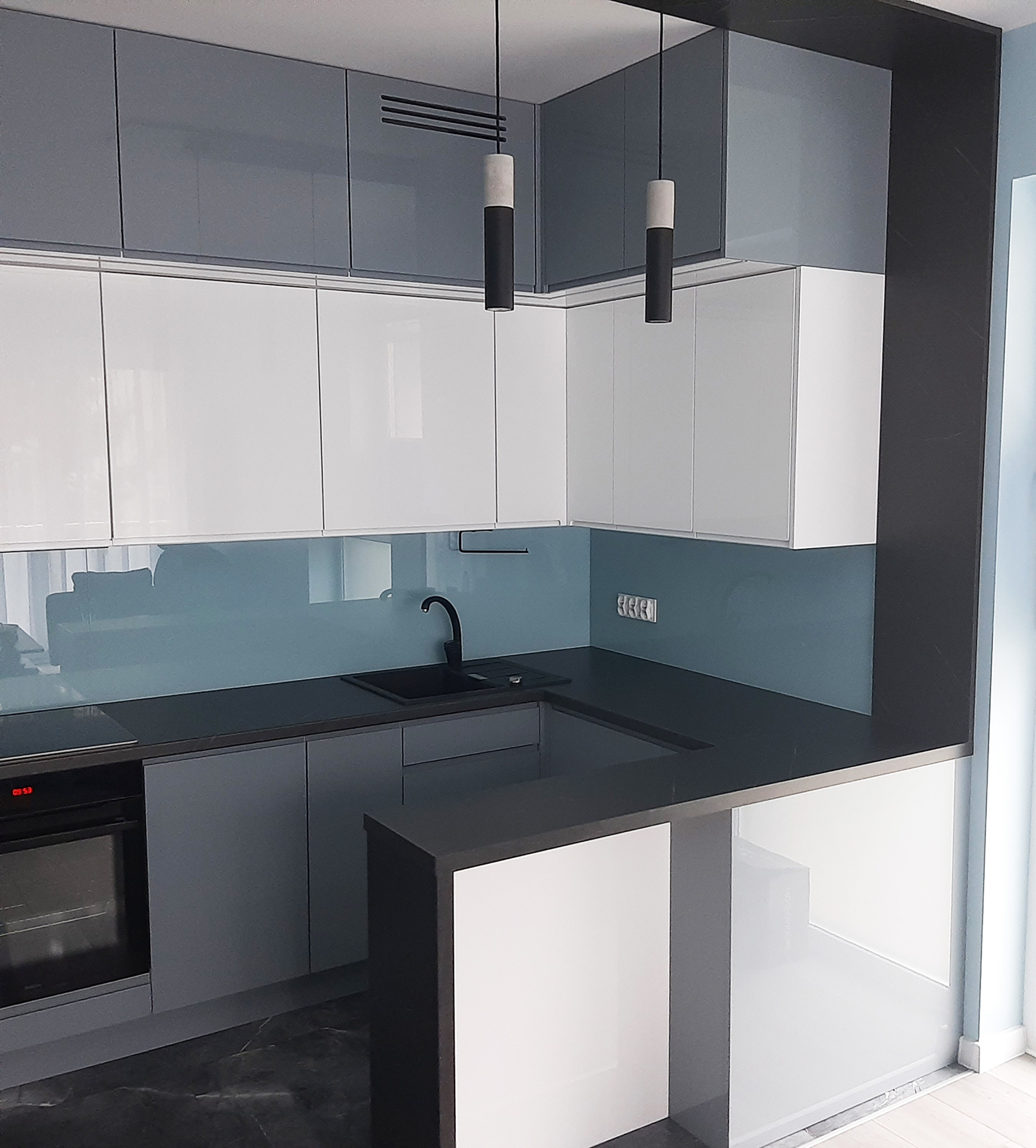 Nowoczesna, narożna kuchnia z szarymi i białymi szafkami z połyskiem, czarnym blatem i niebieskim panelem szklanym między blatem a szafkami wiszącymi, oświetlona dwoma podłużnymi lampami.