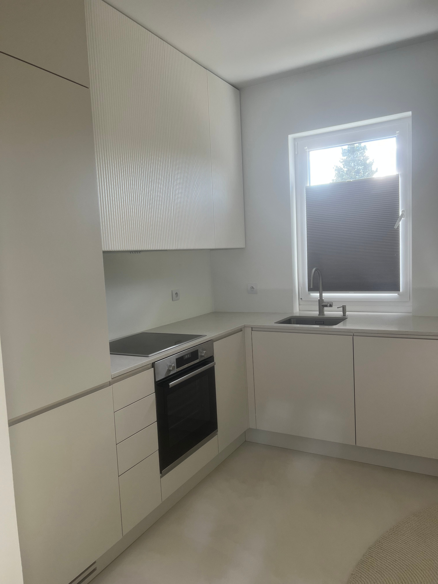 Jasna kuchnia z minimalistycznymi frontami szafek, zintegrowanym sprzętem AGD i żaluzjami plisowanymi w oknie.