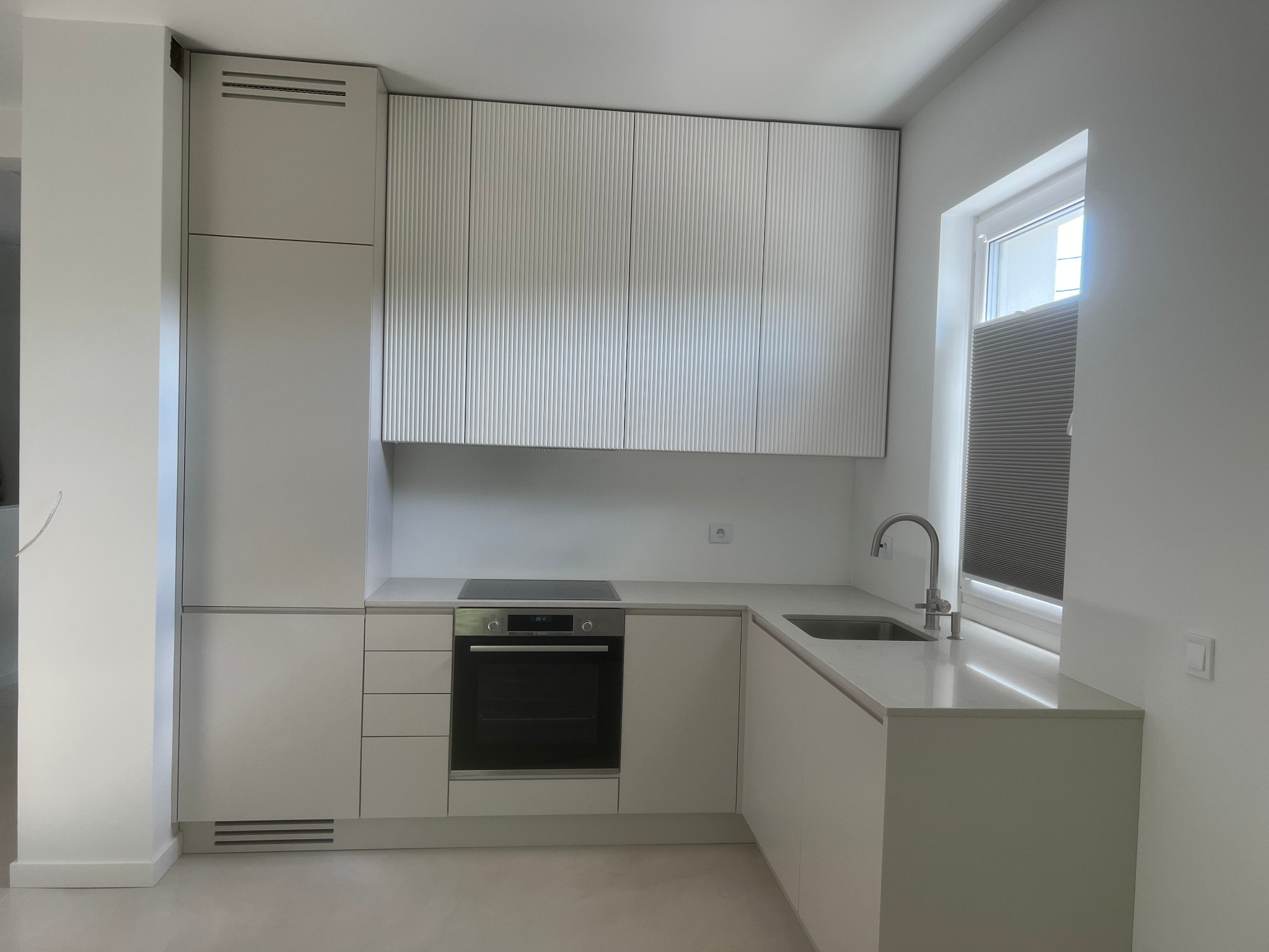 Nowoczesna, minimalistyczna kuchnia w odcieniach bieli z charakterystycznymi ryflowanymi frontami szafek wiszących i wbudowanym piekarnikiem pod płytą grzewczą, oświetlona naturalnym światłem...