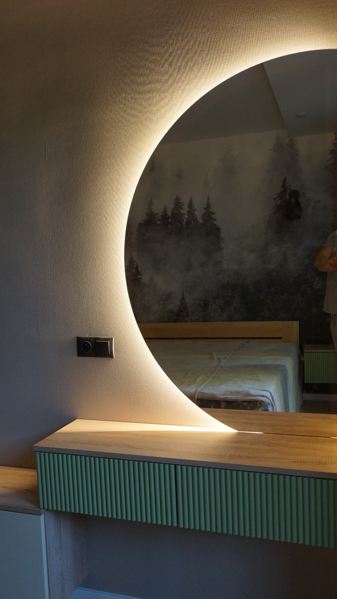 Toaletka z okrągłym lustrem z podświetleniem LED, zielone lamele na frontach szuflad, w tle tapeta z motywem leśnym i fragment łóżka.