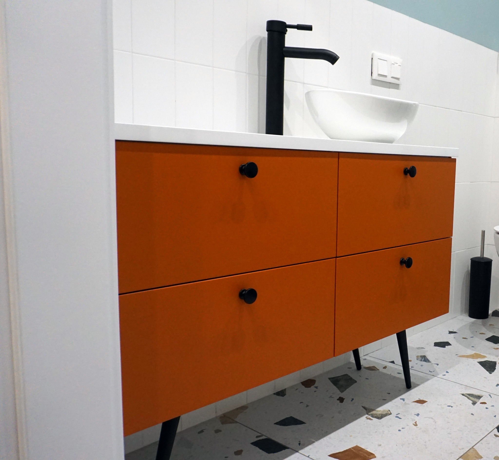 Pomarańczowa szafka łazienkowa z czterema szufladami, czarnymi gałkami i nóżkami, biała umywalka nablatowa i czarna bateria, na tle białych płytek i podłogi w terrazzo.