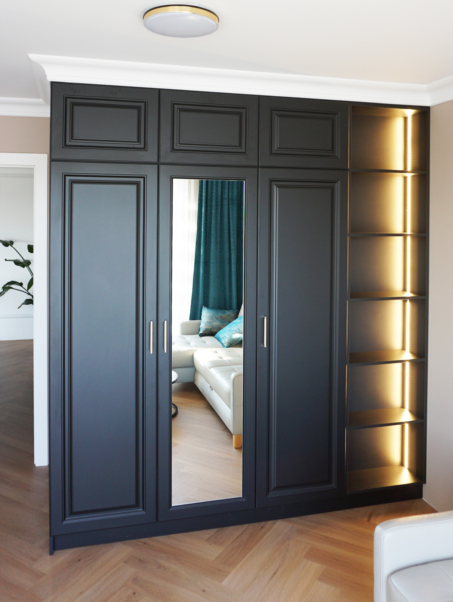 Czarna, zabudowana szafa z lustrem i podświetlanymi półkami, odbijająca fragment salonu z białą sofą i turkusowymi zasłonami.