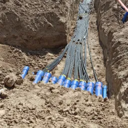 Wykop w ziemi z ułożonymi kablami w niebieskich rurach ochronnych z numeracją, widok z góry.