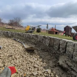 Budowa murku z kostki granitowej: widok na wykop z podsypką, narzędzia (szpadel, młotek, poziomica), ubijarkę i taczkę w tle ogrodu z domkiem zabaw i grillem.