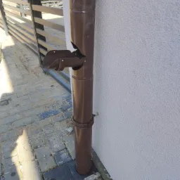 Brązowa rura spustowa z osadnikiem na kostce brukowej, przy jasnej elewacji budynku i metalowej bramie.