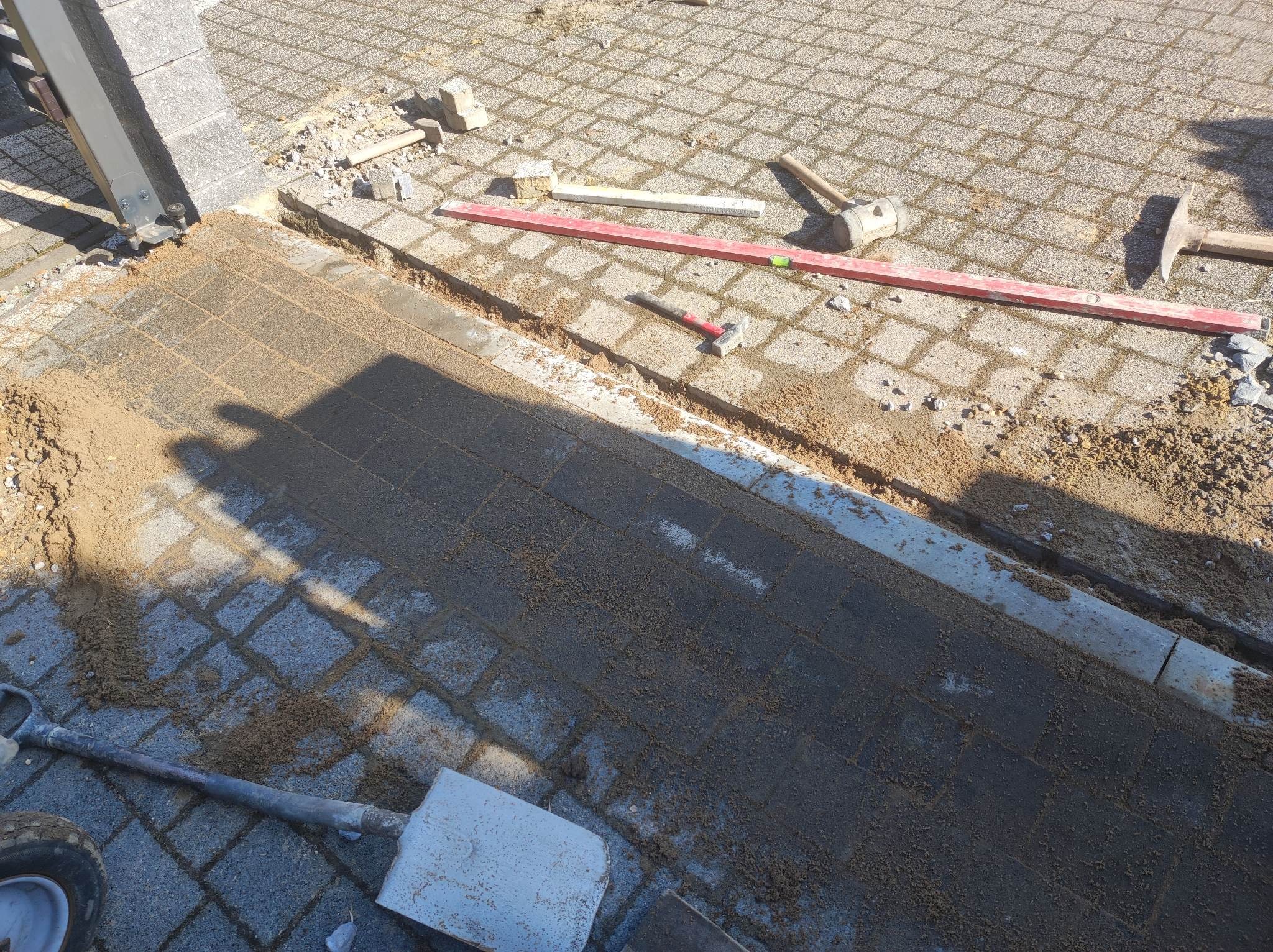 Układanie kostki brukowej: przygotowane podłoże z piaskiem, narzędzia budowlane (poziomica, młotek) widoczne na częściowo ułożonej nawierzchni z kostki.