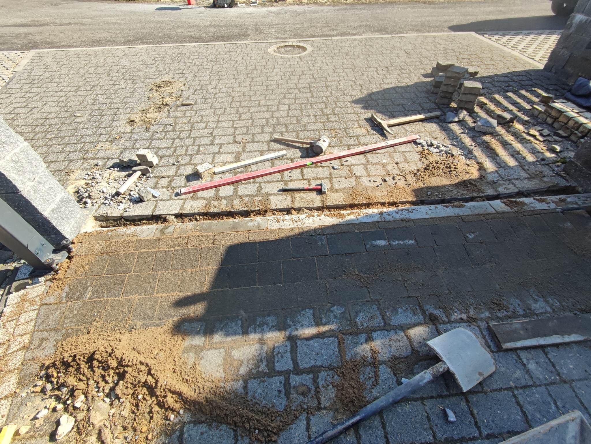 Fragment chodnika z kostki granitowej w trakcie układania, widoczne narzędzia: poziomica, młotek, łopata, oraz stos kostki do ułożenia.