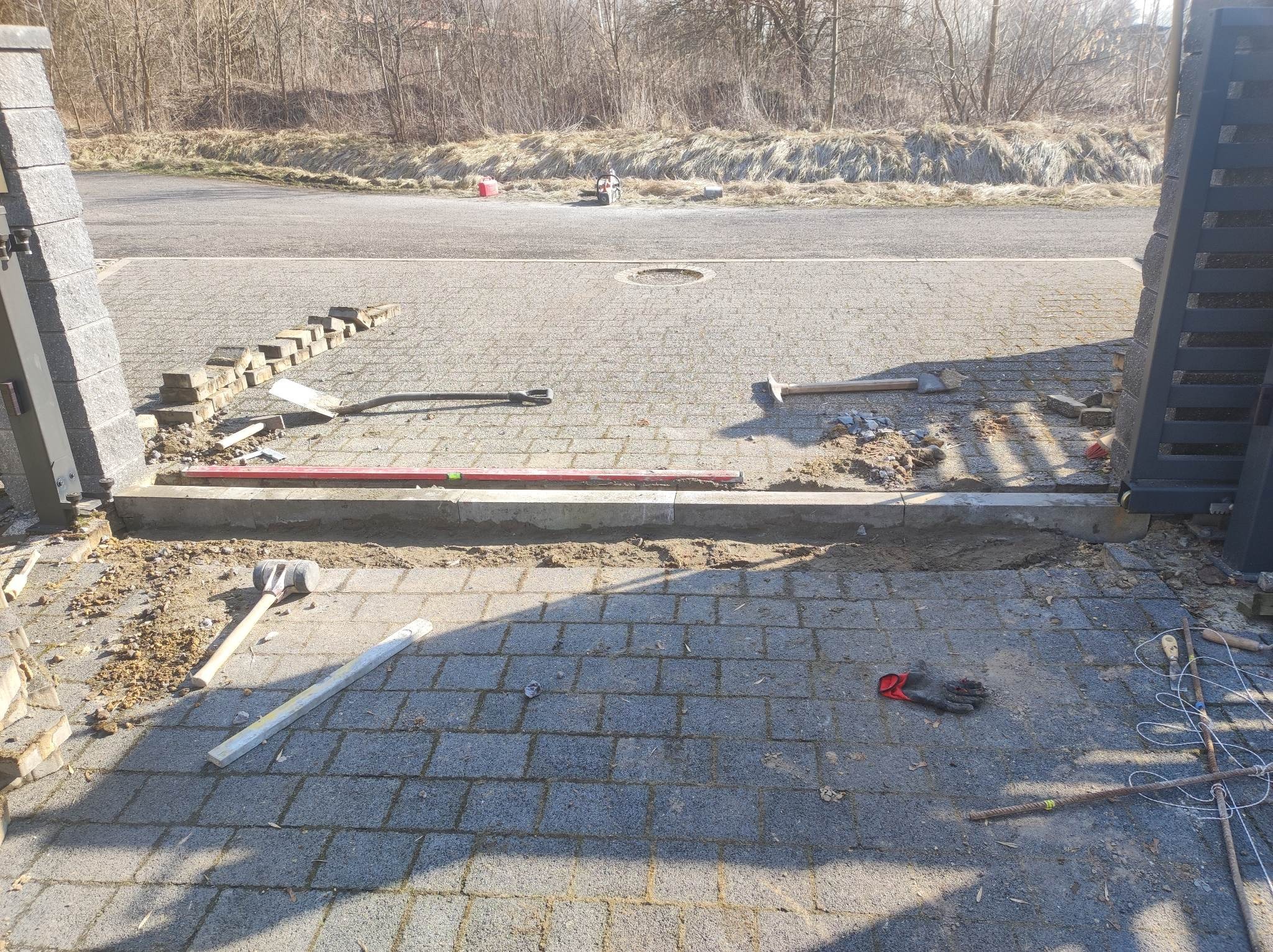 W trakcie prac: ułożona część podjazdu z kostki brukowej przed posesją, widoczne narzędzia, poziomica, gumowy młotek, rękawice robocze oraz przygotowane obrzeża i kostka do dalszego układania.