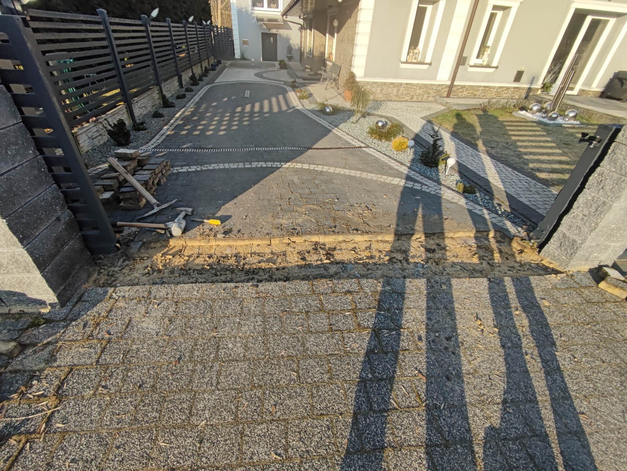 Fragment podjazdu z szarej kostki brukowej w trakcie układania, widoczne narzędzia: młotek i kilof, oraz stos kostki przy ogrodzeniu z poziomych desek. W tle dom z szarą elewacją i dekoracyjnymi...