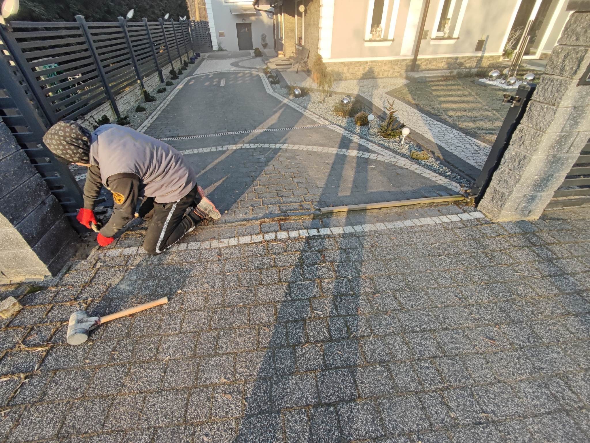 Pracownik w czerwonych rękawicach układa kostkę granitową przy nowym ogrodzeniu, widoczny gumowy młotek i fragment gotowego podjazdu.