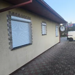 Elewacja budynku z trzema oknami zabezpieczonymi roletami zewnętrznymi. Okna obramowane dekoracyjną imitacją kamienia. Zewnętrzna antena satelitarna na ścianie.