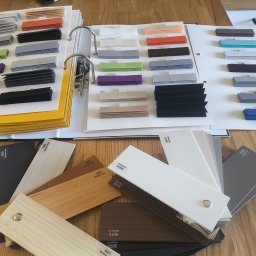 Katalog z próbkami kolorów i materiałów wykończeniowych, w tym drewnopodobnych, ułożony na drewnianym blacie. Widoczne różne odcienie i tekstury.