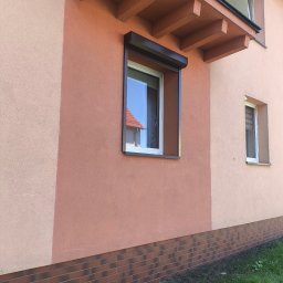 BFS Bartłomiej Faryński - Brązowa roleta zewnętrzna zamontowana na oknie budynku z dwukolorową elewacją i ceglaną podmurówką. Widok z dołu, na pierwszym planie trawa.