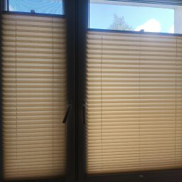 BFS Bartłomiej Faryński - Beżowe rolety plisowane w oknie z widokiem na niebo w Podańsku. Dwie części rolety, widoczne uchwyty okienne, delikatna zasłona po prawej stronie.