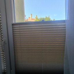 BFS Bartłomiej Faryński - Beżowa roleta plisowana w oknie z widokiem na budynek z czerwonym dachem i drzewa. Biała rama okienna i fragment zasłony po prawej stronie kadru.