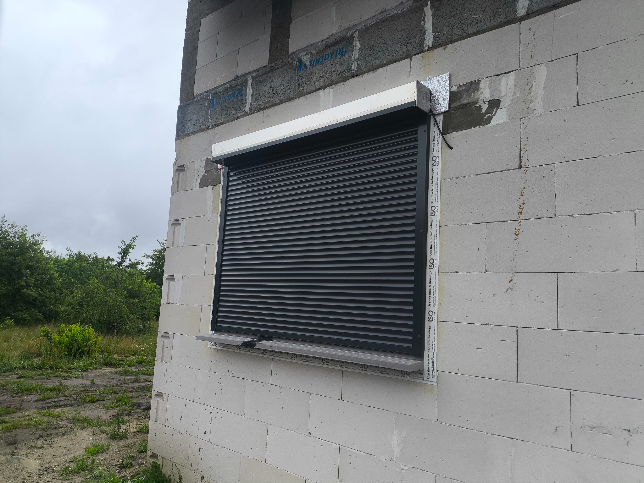 Zamknięta, szara żaluzja zewnętrzna zamontowana w oknie budynku w trakcie budowy, z widocznymi bloczkami betonowymi i zielenią w tle.