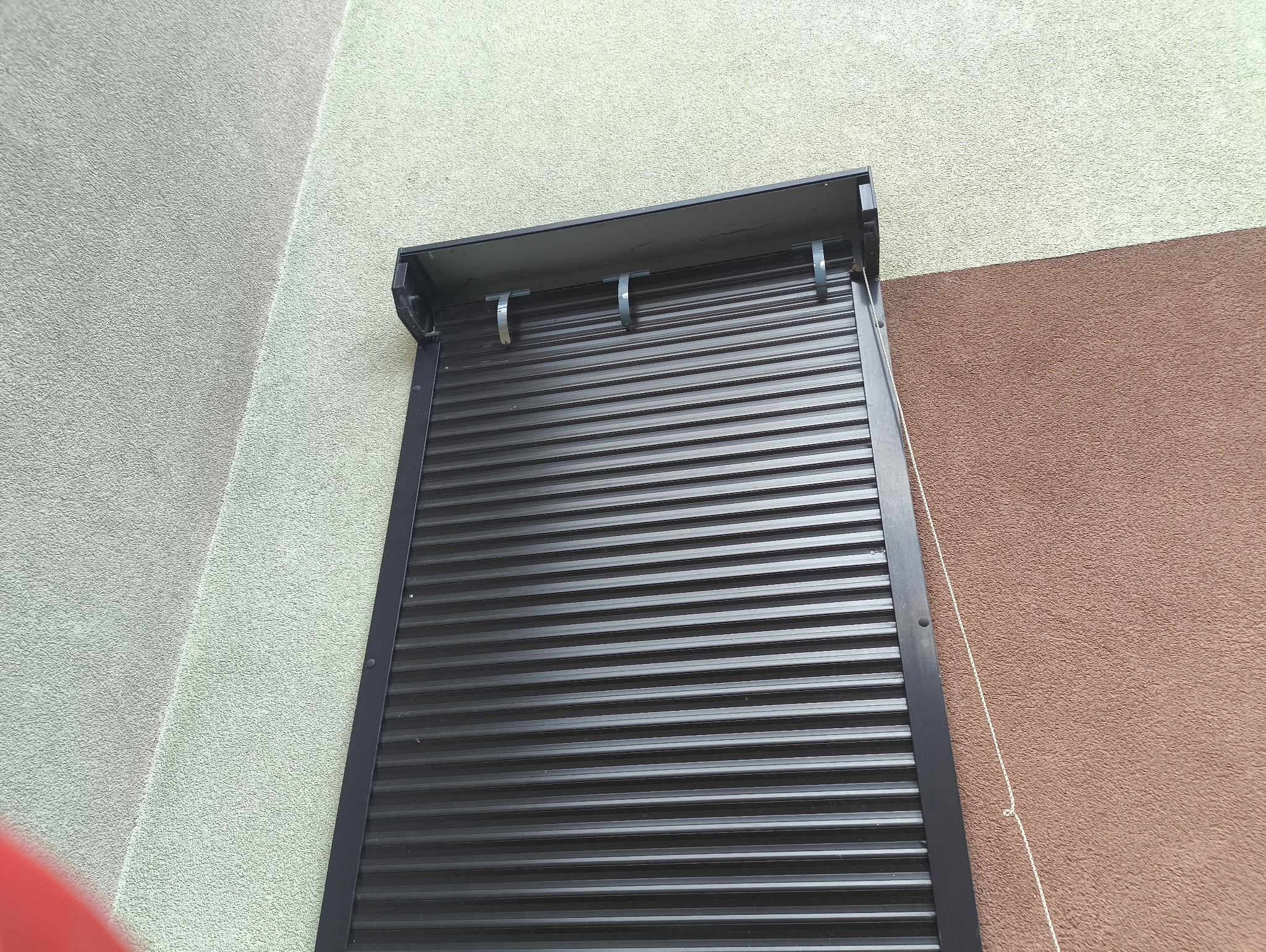 Ciemne, metalowe żaluzje zewnętrzne częściowo zasłonięte, z widocznym mechanizmem zwijania, na tle dwukolorowej elewacji budynku. Ujęcie z dołu.