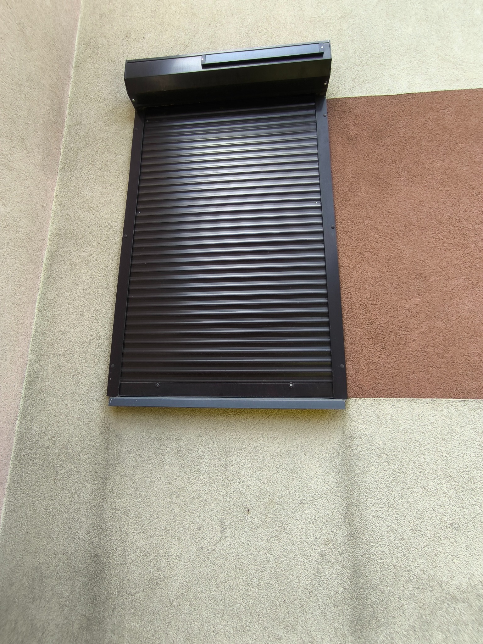 Zamknięta, brązowa roleta zewnętrzna zamontowana na elewacji budynku o dwukolorowej, piaskowo-brązowej strukturze tynku. Widok z dołu, na fasadzie.