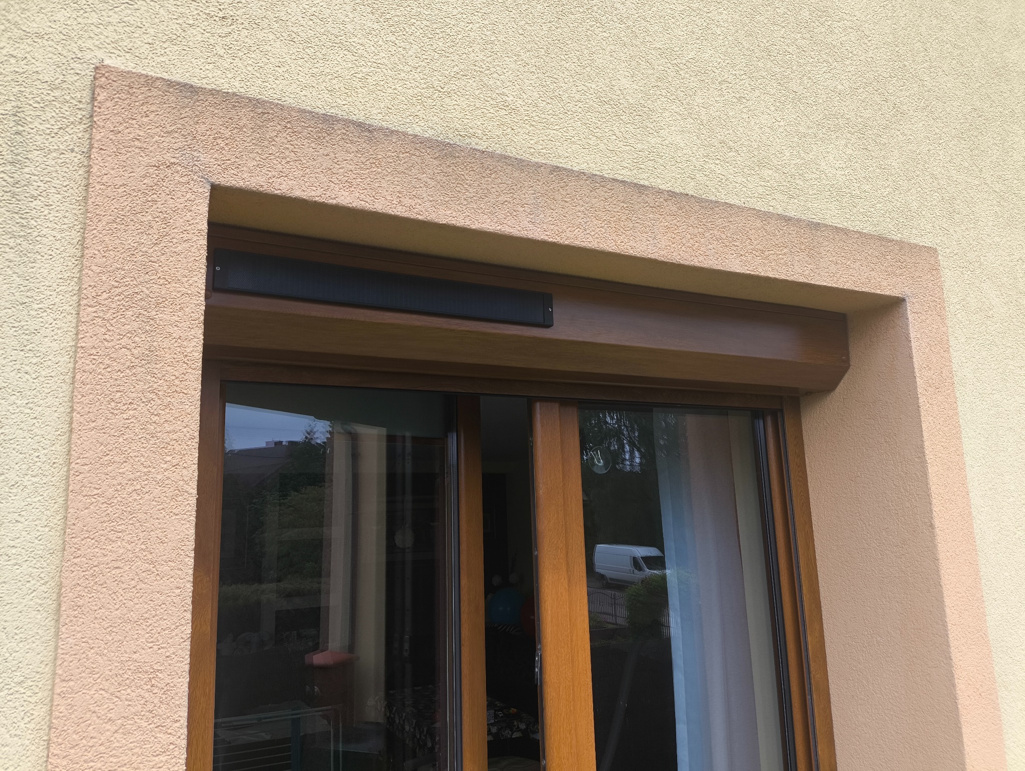 Zamknięta roleta zewnętrzna w brązowej ramie okiennej na tle beżowej elewacji budynku. Widoczny fragment okna z refleksami otoczenia.