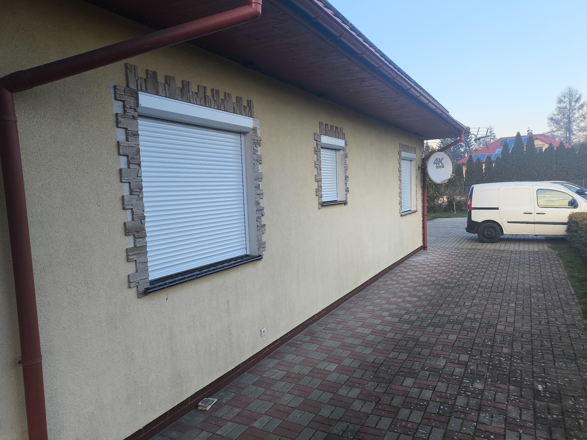 Elewacja budynku z trzema oknami zabezpieczonymi roletami zewnętrznymi. Okna obramowane dekoracyjną imitacją kamienia. Zewnętrzna antena satelitarna na ścianie.