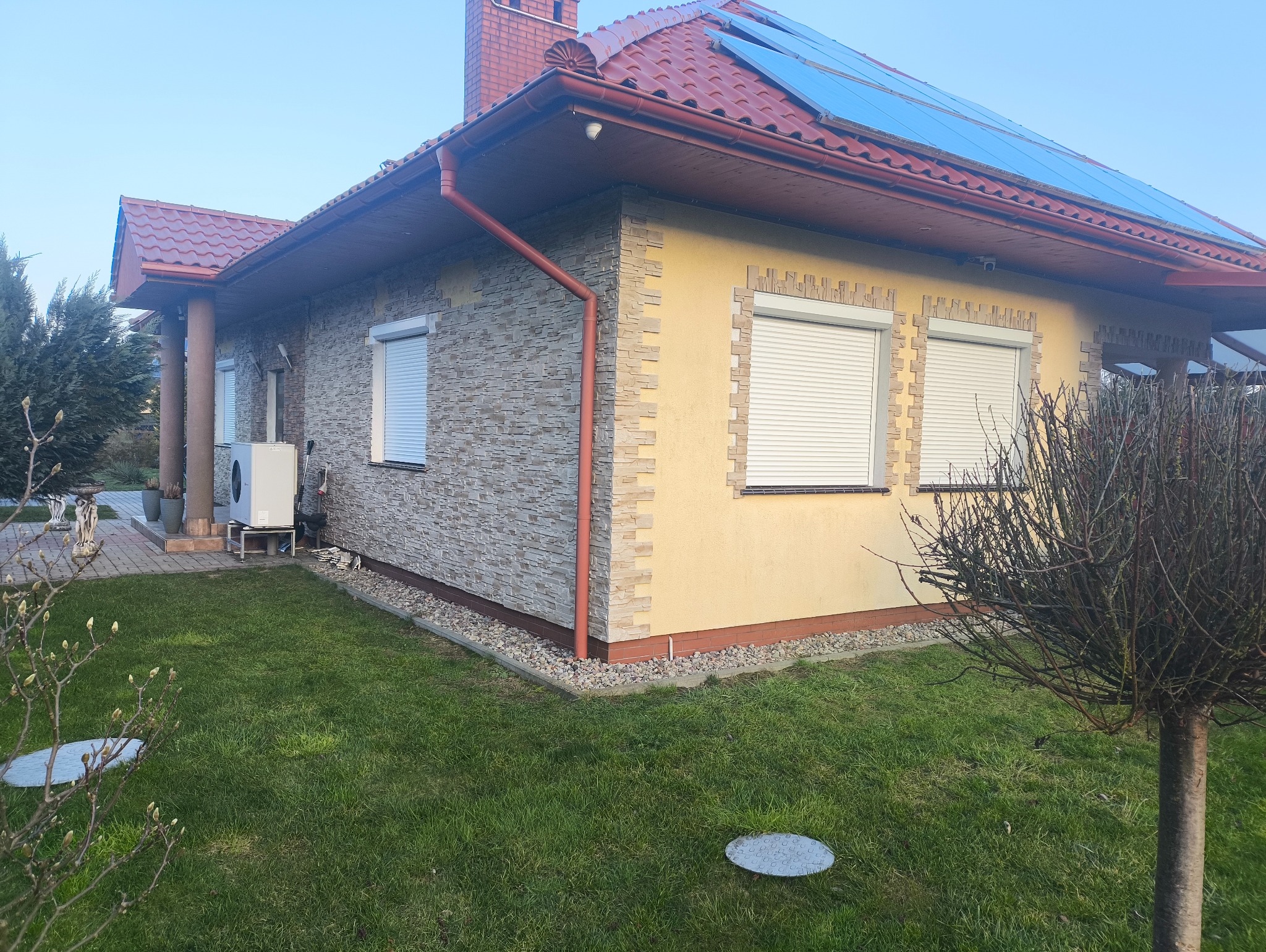 Elewacja domu z kamienną okładziną i żółtym tynkiem, okna z żaluzjami zewnętrznymi. Czerwona dachówka i panele słoneczne na dachu. Widok z zewnątrz na trawnik.