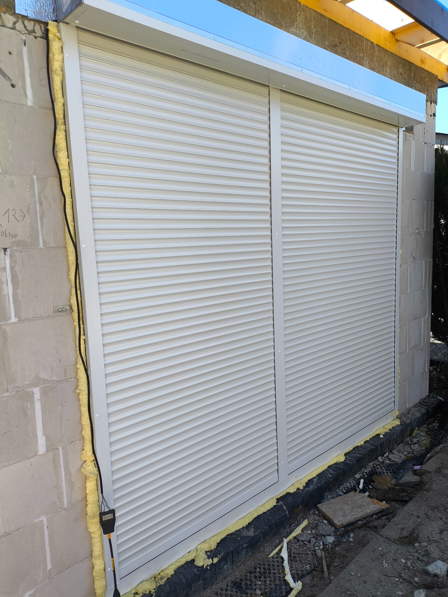 Zamknięte, białe rolety zewnętrzne zamontowane w surowym murze. Widoczne uszczelnienia pianką montażową. Ujęcie z dołu, podkreślające solidność instalacji.
