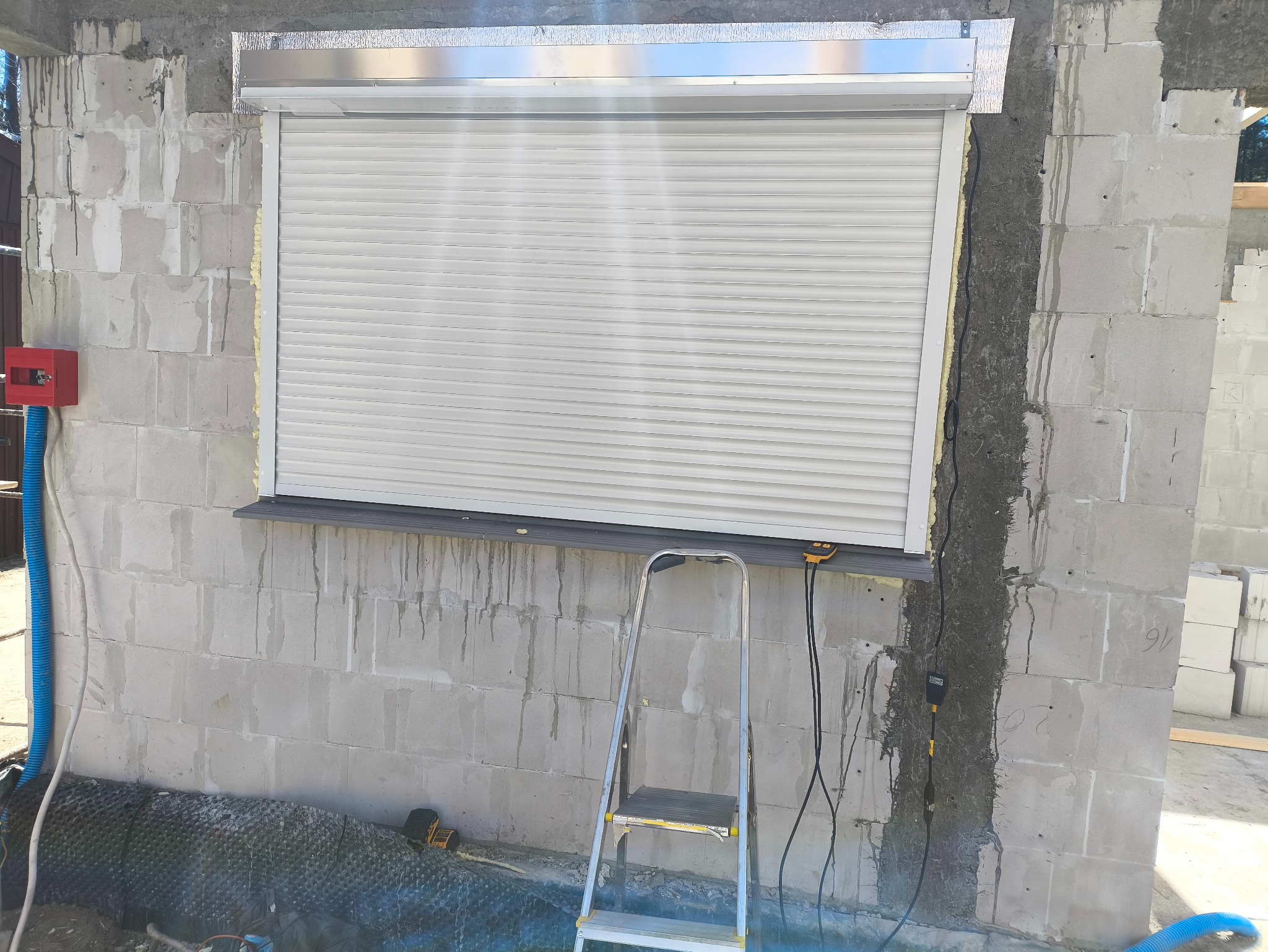Zamknięta, biała roleta zewnętrzna zamontowana w oknie budynku w trakcie budowy. Widoczne bloczki betonowe, drabina i przewody elektryczne.