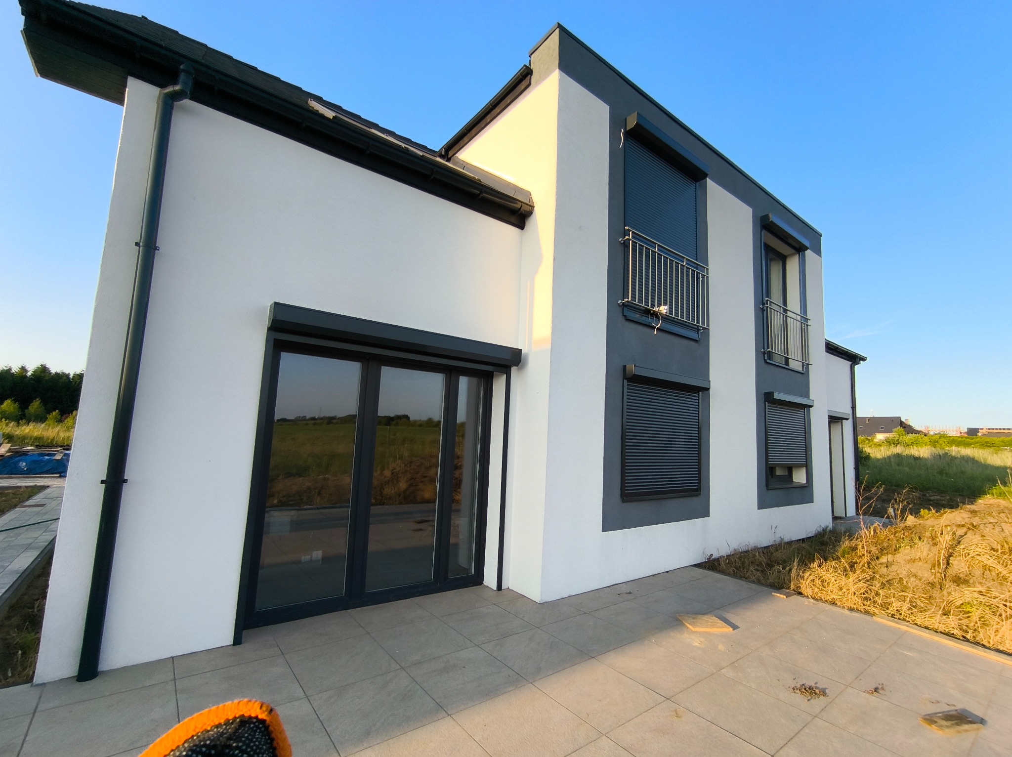 Nowoczesny dom z szarymi roletami zewnętrznymi i balkonem z metalową balustradą, uchwycony w słoneczny dzień z niskiej perspektywy.