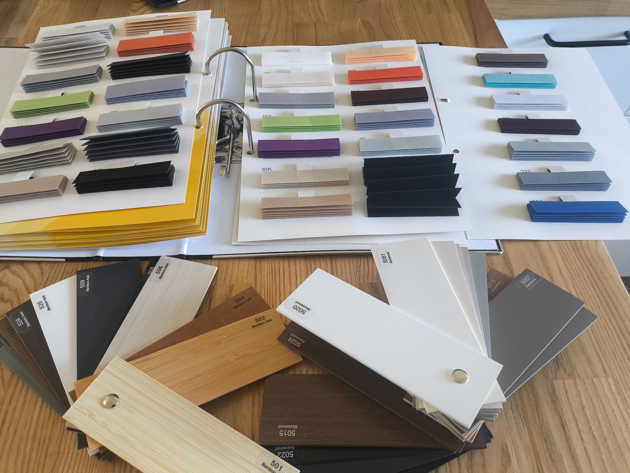 Katalog z próbkami kolorów i materiałów wykończeniowych, w tym drewnopodobnych, ułożony na drewnianym blacie. Widoczne różne odcienie i tekstury.