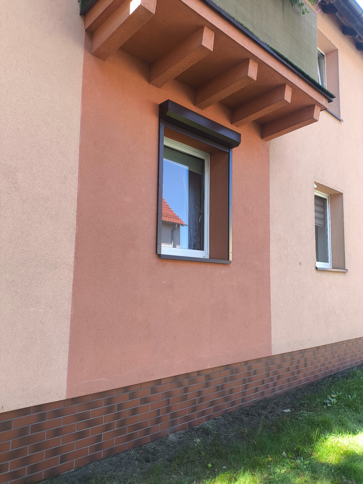 Brązowa roleta zewnętrzna zamontowana na oknie budynku z dwukolorową elewacją i ceglaną podmurówką. Widok z dołu, na pierwszym planie trawa.