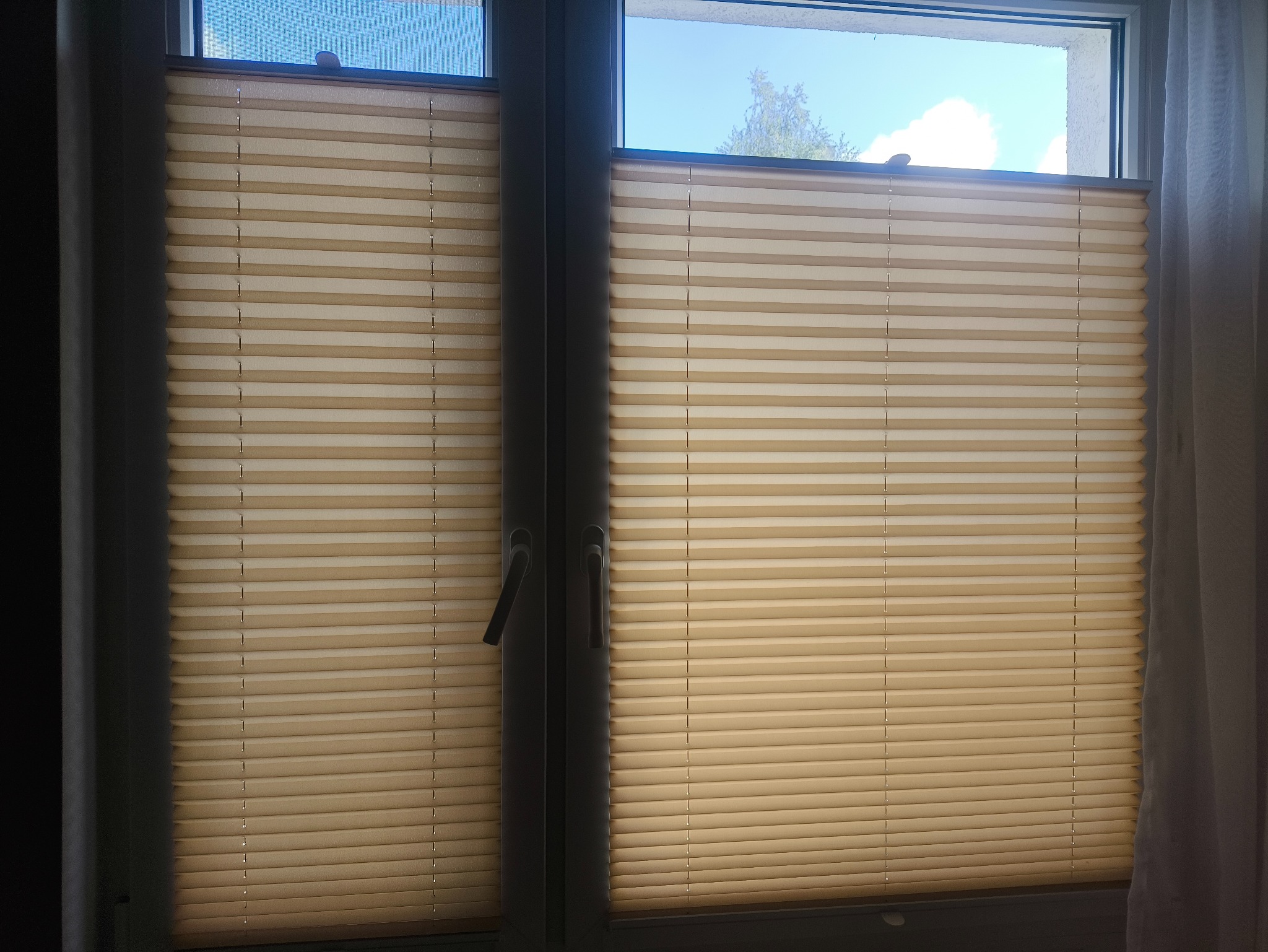 Beżowe rolety plisowane w oknie z widokiem na niebo w Podańsku. Dwie części rolety, widoczne uchwyty okienne, delikatna zasłona po prawej stronie.