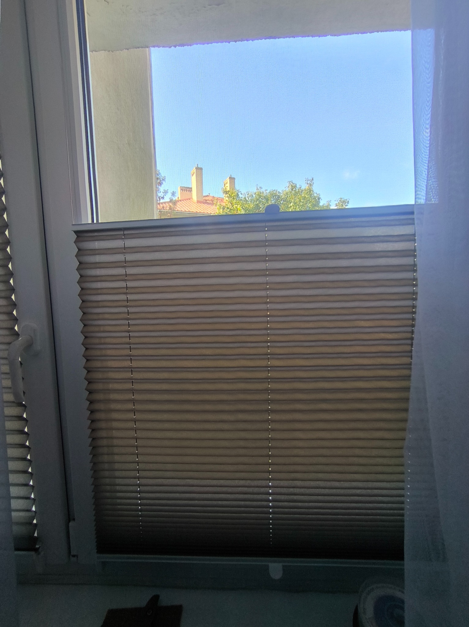 Beżowa roleta plisowana w oknie z widokiem na budynek z czerwonym dachem i drzewa. Biała rama okienna i fragment zasłony po prawej stronie kadru.