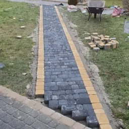 Ścieżka z szarej kostki brukowej z żółtymi obrzeżami w trakcie układania, widoczne niedokończone fragmenty, stos kostki obok, taczka i dom w tle.