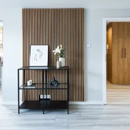 Nowoczesne wnętrze mieszkania z drewnianą ścianką dekoracyjną z pionowych listew, czarnym metalowym regałem z dekoracjami, minimalistycznym obrazem i białymi liliami w wazonie, widok na kuchnię...