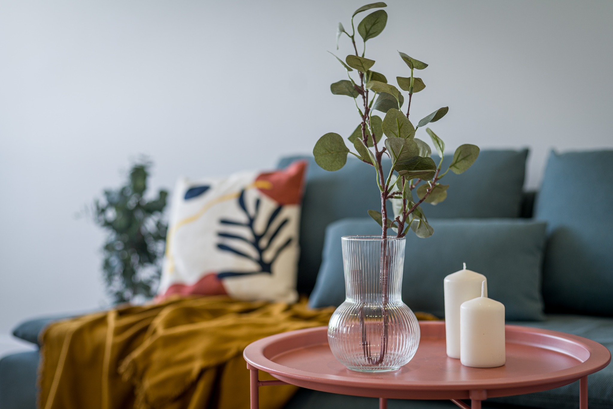 Dekoracyjny stolik kawowy z różowym blatem, na którym znajdują się szklany wazon z gałązkami eukaliptusa i dwie białe świece, w tle turkusowa sofa z poduszkami i musztardowy pled.